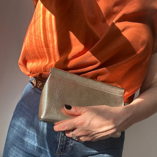 Person holding a gold clutch with an orange top and blue jeans against a neutral background