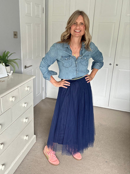 A woman stands in a bedroom wearing a light blue denim shirt tucked into a long navy blue tulle skirt, paired with pink trainers. She is positioned in front of white closet doors and a white dresser.