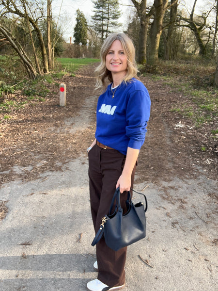 woman in brown trousers and cobalt blue top holding a navy leather bag and standing in a forest