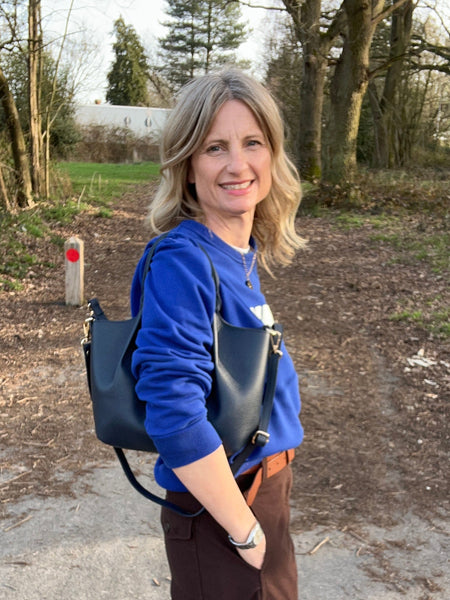 woman in brown trousers and cobalt blue top carrying a navy leather bag and standing in a forest