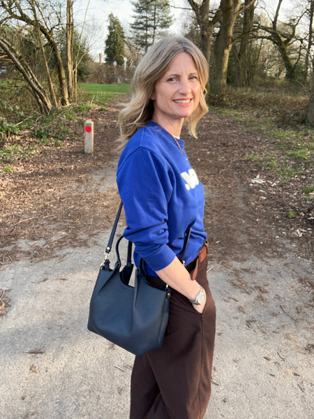 woman in brown trousers and cobalt blue top wearing a navy leather bag and standing in a forest