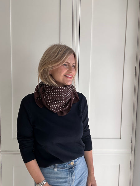 A woman with blonde hair wearing a dark brown infinity scarf with white polka dots, a blue long-sleeve top, and light blue jeans, standing in front of white paneled doors.