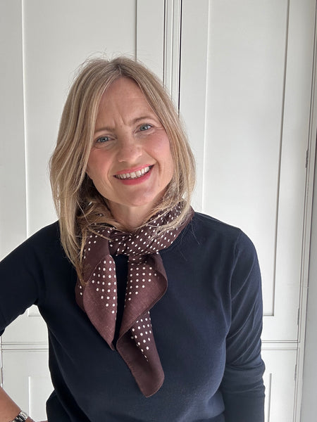 A smiling blonde woman wearing a dark navy long-sleeve top and a brown silk scarf with white polka dots, standing in front of a white paneled door.