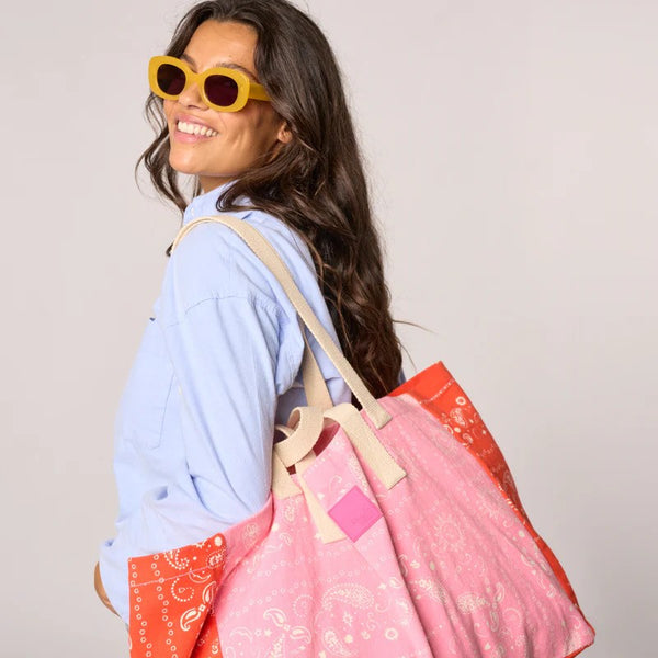 Woman wearing yellow sunglasses and holding a pink and orange patterned bag against a plain background
