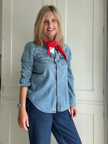 Woman wearing a denim shirt and blue jeans with a red scarf, standing indoors.