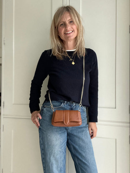 Woman wearing a black top and blue jeans, holding a brown leather bag indoors.