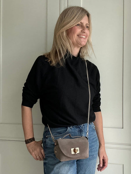 Woman wearing a black top and blue jeans, wearing a taupe handbag indoors.
